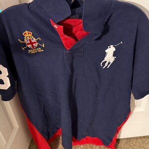 May include: A navy blue short-sleeved polo shirt with a red inner placket and red side panels. The chest features a white embroidered polo player logo and a crest with the text MMIX MERCER POLO TEAM. A large white number 3 is on the sleeve.