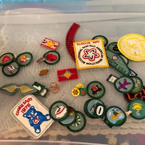 Op de afbeelding: Een verzameling Girl Scout-patches, waaronder een "Cookie Sales 1976" patch met een blauwe beer en een "Girl Scouts USA" patch. Andere patches hebben verschillende ontwerpen en tekst, in een doorzichtige plastic container.