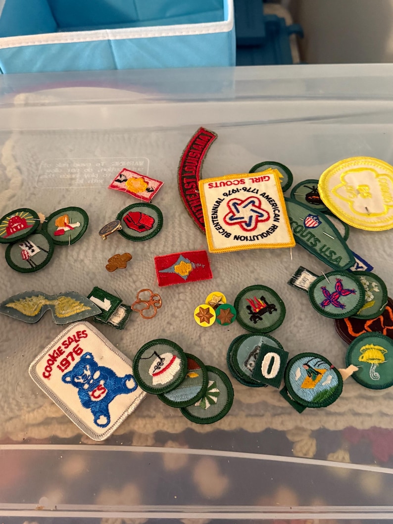 Op de afbeelding: Een verzameling Girl Scout-patches, waaronder een "Cookie Sales 1976" patch met een blauwe beer en een "Girl Scouts USA" patch. Andere patches hebben verschillende ontwerpen en tekst, in een doorzichtige plastic container.