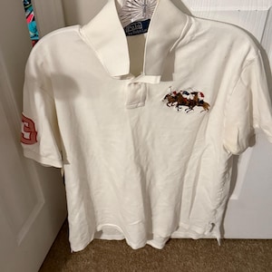 May include: A white short-sleeved polo shirt featuring a collar and a button placket. The chest displays an embroidered graphic of polo players on horses. A large red number three is embroidered on the right sleeve. The label reads Polo by Ralph Lauren.