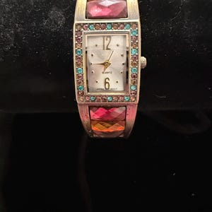 May include: A rectangular quartz watch with a silver-tone metal band featuring faceted, multi-colored stones. The watch face is white with gold-tone hands, the numbers 12 and 6, and the text QUARTZ and JAPAN MOVT. The bezel is lined with small, colorful crystals.