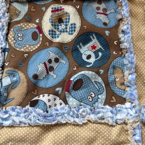 Peut inclure: Une courtepointe en patchwork avec un carré central sur fond marron et des motifs circulaires bleus illustrant des chiens. La courtepointe présente des coutures effilochées à carreaux bleus et blancs et un tissu beige à pois blancs.