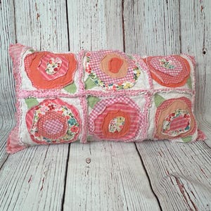May include: A rectangular decorative pillow featuring a patchwork design with six squares. Each square showcases a fabric flower in shades of pink, coral, and white. The pillow has a ruffled edge and is set against a light wood background.