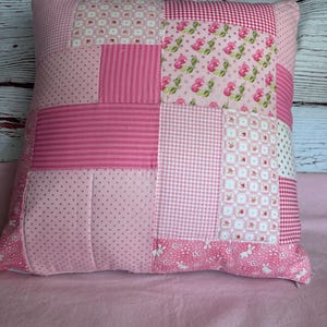 May include: A square patchwork pillow featuring various shades of pink fabric. The design includes panels with patterns such as small floral prints, vertical stripes, pink and white gingham, polka dots, and a repeating white rabbit motif on a pink background.
