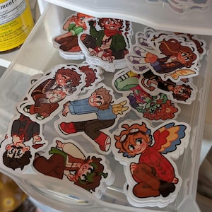 May include: A collection of colorful stickers featuring cartoon characters in various poses. The stickers showcase characters with wings, holding animals, and wearing different outfits. The stickers are in a clear plastic container.