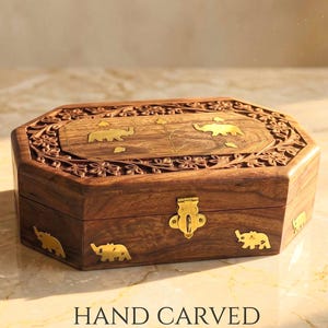 Può includere: Una scatola ottagonale in legno con intagli floreali complessi e motivi di elefanti in ottone intarsiati sul coperchio e sui lati. La scatola presenta un meccanismo di chiusura centrale in ottone sul davanti e una finitura in legno scuro.