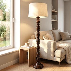 Può includere: Una lampada da terra con base in legno tornito marrone scuro e paralume cilindrico in tessuto color bianco sporco. La lampada poggia su una base circolare e presenta una serie di segmenti in legno arrotondati impilati lungo la colonna.