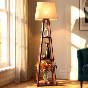 Op de afbeelding: Een hoge, donkerbruine houten vloerlamp met een piramidevormig frame met drie geïntegreerde planken. De bovenste plank bevat een witte keramische vaas, de middelste plank toont een zwart beeldhouwwerk en de onderste plank bevat een glazen vaas met oranje bloemen.
