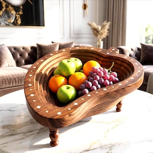 Op de afbeelding: Een houten fruitschaal met een uniek, gebogen ontwerp, gevuld met vers fruit. De schaal is gemaakt van donker hout en heeft ingelegde witte accenten. De schaal bevat groene appels, sinaasappels en een tros paarse druiven. De schaal staat op een marmeren tafel.