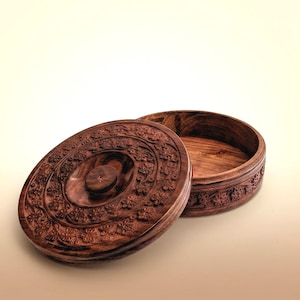 Puede incluir: Una caja redonda de madera con tapa, con intrincadas tallas florales. La caja es de madera marrón oscuro, con la tapa ligeramente entreabierta, revelando el interior. La tapa tiene un pomo central y patrones florales detallados.