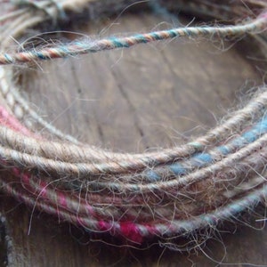 Op de afbeelding: Een close-up van een bol garen met een natuurlijke bruine kleur en dunne strengen roze, blauwe en witte garen die door het hele garen zijn geweven.