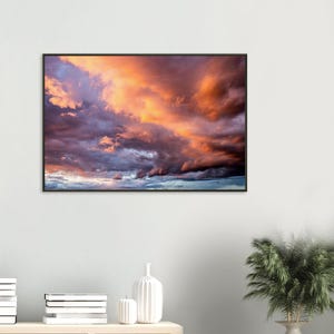 May include: A rectangular wall art print featuring a dramatic sky filled with dense, layered clouds. The scene displays a vibrant sunset color palette, transitioning from deep purples and dark blues in the lower sections to bright orange and gold hues.