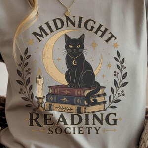 以下が含まれることがあります： スポーツグレーのTシャツ。三日月を背景に本の上に座る黒猫のグラフィック。テキストは「MIDNIGHT READING SOCIETY」と書かれています。デザインにはろうそくと星が含まれています。