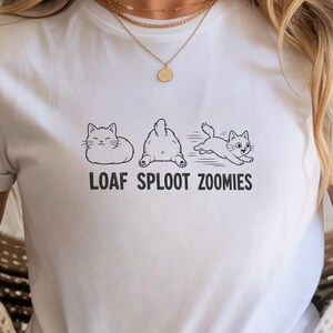 Może przedstawiać: Biała koszulka z czarną grafiką przedstawiającą trzy koty w różnych pozach, z napisem "LOAF SPLOOT ZOOMIES" poniżej. Koszulka wykonana jest z miękkiego materiału.