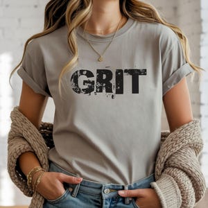 May include: A gray t-shirt with the word "GRIT" printed in distressed black lettering. The shirt has short sleeves and is paired with blue jeans. A beige sweater is draped over the shoulders.