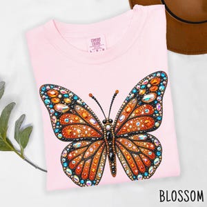 May include: A light pink t-shirt featuring a large butterfly graphic embellished with orange, blue, and clear faceted rhinestones. The shirt has a Comfort Colors brand label at the inner neckline with size L-G-G printed on the tag.