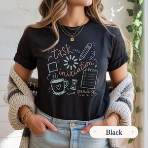 May include: A black short-sleeve t-shirt featuring a graphic print with the text "task initiation" and "pending". The design includes a pencil, a notepad, a coffee mug with a heart, an hourglass, a loading icon, and a square outline in light blue and tan.