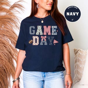 以下が含まれることがあります： ネイビーブルーの半袖Tシャツ。チェック柄、花柄、ギンガムチェックなどの様々なパターンがあしらわれたブロック体の「GAME DAY」という文字がプリントされています。小さなフットボール、ピンクのリボン、「GO TEAM」と書かれたペナント旗のデザインが含まれています。