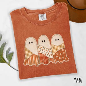 Op de afbeelding: Een kortmouwig T-shirt in gebrand oranje met een centraal applicatieontwerp van drie geesten. Elke geest is opgebouwd uit stoffen patches met diverse patronen, waaronder gingham, ruitjes en bloemenprints, met kleine zwarte ronde ogen.