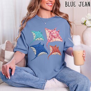 Könnte beinhalten: Ein hellblaues Kurzarm-T-Shirt mit einem Grafikdruck von vier Stachelrochen. Jeder Rochen hat ein eigenes Muster: einer ist blaugrün kariert, einer ist rosa geblümt, einer ist marineblau mit weißen Punkten und einer ist orange-lila gestreift.