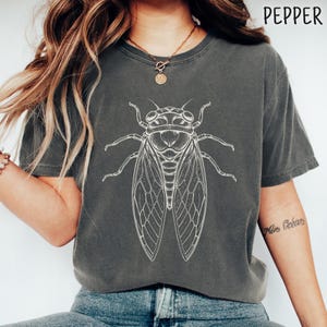 Può includere: Una t-shirt a maniche corte color grigio antracite con un'illustrazione a linee bianche di una cicala centrata sul petto. La maglietta presenta un girocollo e una vestibilità rilassata. La parola PEPPER è stampata nell'angolo in alto a destra.