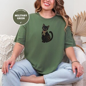 Può includere: Una t-shirt a maniche corte color verde militare con la grafica di un gatto nero che indossa un fiocco beige al collo. Il gatto presenta intricati motivi dorati sul corpo. Un badge circolare in alto a sinistra riporta il testo GILDAN 5000 MILITARY GREEN.
