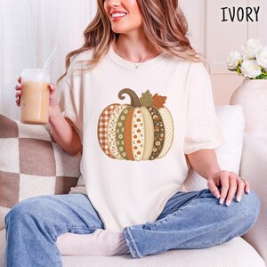 Puede incluir: Una camiseta de manga corta color marfil con un gran gráfico de una calabaza compuesta por paneles verticales con varios patrones, incluyendo cuadros, flores y lunares en tonos naranja, verde, marrón y crema. El texto IVORY es visible.