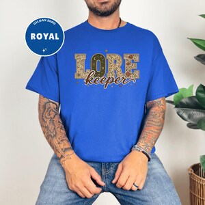 May include: A royal blue short-sleeve t-shirt featuring the text "LORE keeper" in a decorative font. The letters are filled with intricate patterns including celestial motifs, floral designs, and vintage map elements in shades of brown and gold.