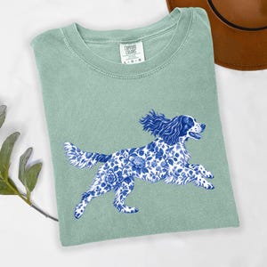 May include: A sage green short-sleeve t-shirt featuring a graphic of a running dog with a blue and white floral pattern. The shirt has a crew neckline and a Comfort Colors brand label printed on the inside of the collar area at the neck.