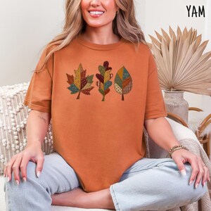 May include: A burnt orange short-sleeve t-shirt featuring a graphic of three autumn leaves. Each leaf is composed of a patchwork of various fabric patterns in shades of green, red, yellow, and brown. The shirt has a crew neckline and a relaxed fit.