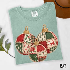 以下が含まれることがあります： セージグリーンのTシャツで、パッチワークデザインの3つのクリスマスオーナメントのグラフィックが描かれています。オーナメントには、赤いトナカイ、緑の松の木、雪の結晶、ヒイラギの実の模様があしらわれています。TシャツにはComfort Colorsのブランドラベルが付いています。