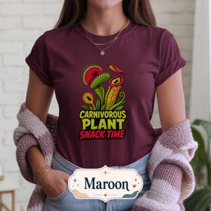 May include: Maroon t-shirt with a vibrant graphic of carnivorous plants, including a Venus flytrap and a pitcher plant, and the text "CARNIVOROUS PLANT SNACK TIME". The text is in yellow and green. The shirt is a solid maroon colour.