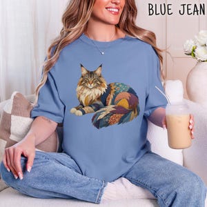 Può includere: Una t-shirt azzurra con la grafica di un gatto a pelo lungo con un motivo patchwork multicolore sul pelo. Il gatto è raffigurato in posizione di riposo con la coda avvolta attorno al corpo. La maglietta presenta un girocollo e maniche corte.