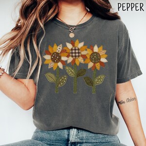 Może przedstawiać: Ciemnoszary t-shirt z krótkim rękawem z grafiką przedstawiającą trzy słoneczniki o płatkach w stylu patchworkowym w odcieniach żółci, pomarańczu i brązu. W prawym górnym rogu znajduje się napis PEPPER wykonany prostą, czarną czcionką bezszeryfową.