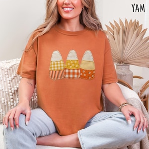 May include: A burnt orange short-sleeve t-shirt featuring a graphic of three candy corn shapes. Each candy corn is designed with a patchwork-style pattern, including gingham, floral, and striped prints in shades of yellow, orange, and white.