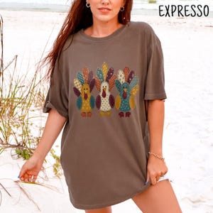 Könnte beinhalten: Ein braunes Kurzarm-T-Shirt mit einem Grafikdruck von drei Truthähnen mit bunten, gemusterten Federn im Patchwork-Stil in Blau-, Gelb-, Orange- und Rottönen. Das Wort EXPRESSO ist in Schwarz in der oberen rechten Ecke aufgedruckt.
