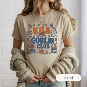 May include: A sand-colored t-shirt featuring a graphic print with the text KILN GOBLIN CLUB in orange and blue. The design includes illustrations of pottery, a kiln, a small creature, and sculpting tools arranged in a decorative layout.
