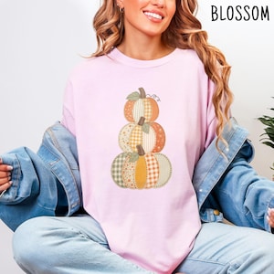 Op de afbeelding: Een lichtroze T-shirt met ronde hals en een afbeelding van drie gestapelde pompoenen. De pompoenen hebben een patchworkpatroon in tinten oranje, geel, groen en wit, inclusief ruitjes- en bloemenprints. Het woord BLOSSOM is afgedrukt.