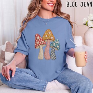 May include: A blue short-sleeve t-shirt featuring a graphic of three mushrooms with different patterns including red gingham, yellow floral, and blue checkered prints. The mushrooms are decorated with small white flower accents.