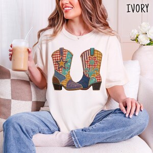 May include: An ivory short-sleeve t-shirt featuring a graphic print of two cowboy boots with a patchwork design. The boots are decorated with red and white gingham, blue floral patterns, and yellow accents, including a star and a flower motif.