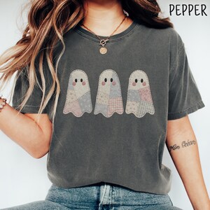 Könnte beinhalten: Ein anthrazitfarbenes Kurzarm-T-Shirt mit einer Grafik von drei Geistern in Patchwork-Mustern in gedeckten Rosa- und Blautönen. Der Text PEPPER ist in der oberen rechten Ecke aufgedruckt. Das T-Shirt hat einen Rundhalsausschnitt.
