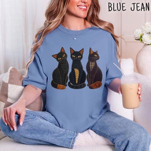 Può includere: Una t-shirt blu a maniche corte con una grafica di tre gatti neri con corpi a motivi geometrici nei toni del nero, marrone e giallo. I gatti sono raffigurati in posizione seduta. La t-shirt è indossata con un paio di jeans blu.