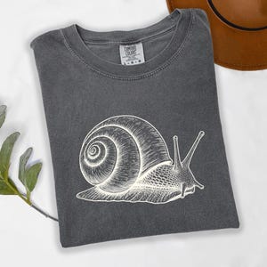 May include: A charcoal gray short-sleeve t-shirt featuring a white line-art illustration of a snail on the front. The shirt has a crew neckline and a Comfort Colors brand label printed on the inside collar with the size marked as L.
