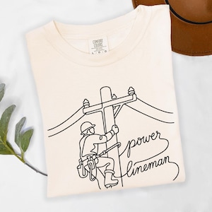 May include: A cream-colored t-shirt featuring a black line art graphic of a utility worker on a power pole. The design includes the text "power lineman" in a cursive script. The shirt has a Comfort Colors brand label indicating a size large.