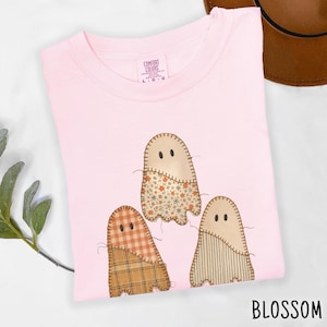 May include: A light pink t-shirt featuring a graphic of three ghosts with stitched edges. The ghosts have patterned lower sections in floral, plaid, and striped designs. The inner neck label reads Comfort Colors with a size L.