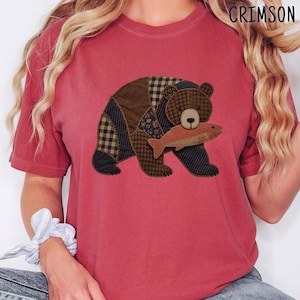 May include: A faded red short-sleeve t-shirt featuring a graphic of a bear made of various brown and black fabric patches, including plaid and floral patterns. The bear is holding a brown fish in its mouth. The word CRIMSON is in the corner.