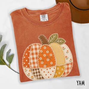 May include: A rust-coloured t-shirt featuring a large pumpkin applique made of various fabric patches including gingham, floral, and striped patterns. The shirt has a Comfort Colors label at the neck and is sized as a large.