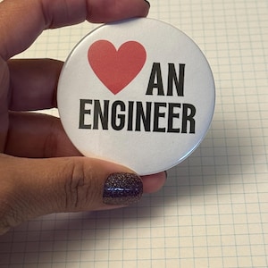 May include: A white round button with a red heart and the words "I LOVE AN ENGINEER" in black. The button is held in a hand.