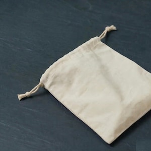 Puede incluir: Una bolsa de cordón de color crema hecha de una tela suave y natural. La bolsa es rectangular con una parte superior redondeada y un cierre de cordón. El fondo es una superficie oscura y texturizada.