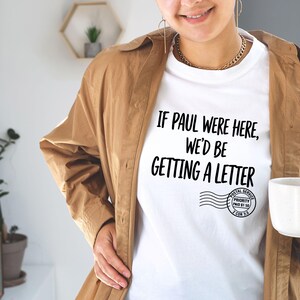 Puede incluir: Camiseta blanca con el texto "IF PAUL WERE HERE, WE'D BE GETTING A LETTER" en negro. La camiseta tambi&eacute;n presenta un gr&aacute;fico de sello postal. La persona lleva una chaqueta marr&oacute;n y sostiene una taza blanca.